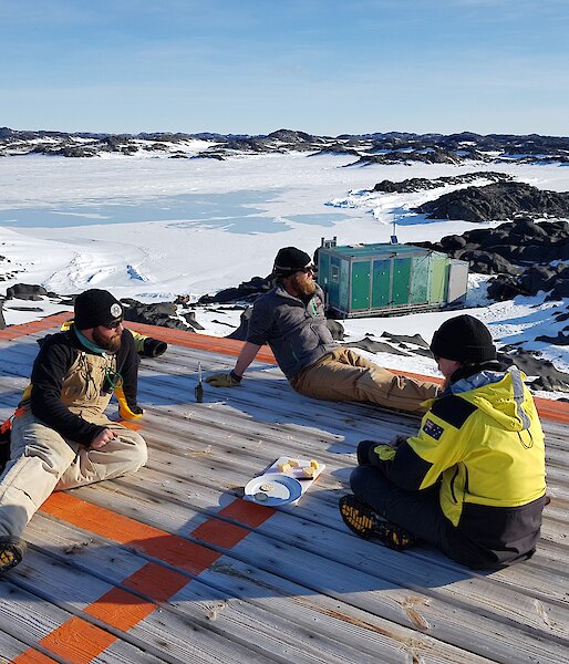 Three expeditioners sit relaxing on the wooden verandah of the hut with cheese and buscuits