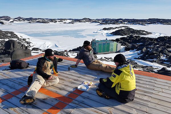 Three expeditioners sit relaxing on the wooden verandah of the hut with cheese and buscuits