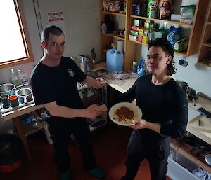 Two expeditioners in the kitchen.  One holding a plate of food and the other a whisk.  They are shaking hands as one presents the whisk to the other.