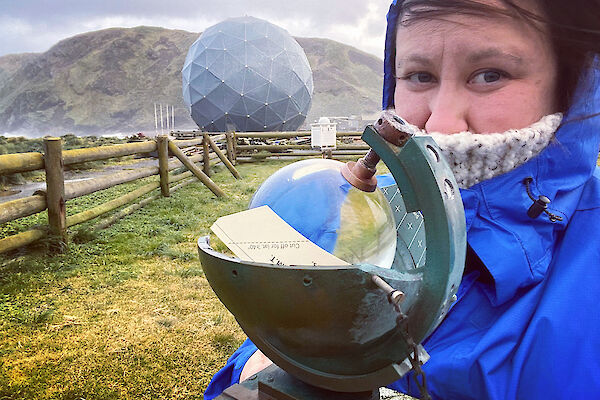Alana standing next to a glass globe sunshine recorder, with the ANARE satellite in the background