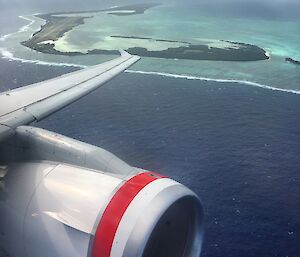 View out an airplane window towards a tropical island and lagoon surrounded by ocean.