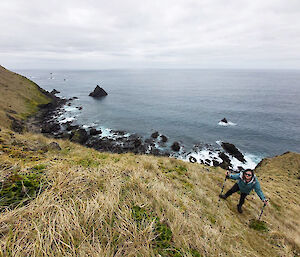 Alana walking up a grassy tussocky hill with the ocean down below