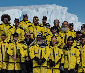 A formal group shot of expeditioners, all in yellow AAD gear, in front of an iceberg