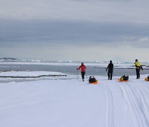 Three expeditioners hauling pack sleds over the snow. Icebergs in the distance.