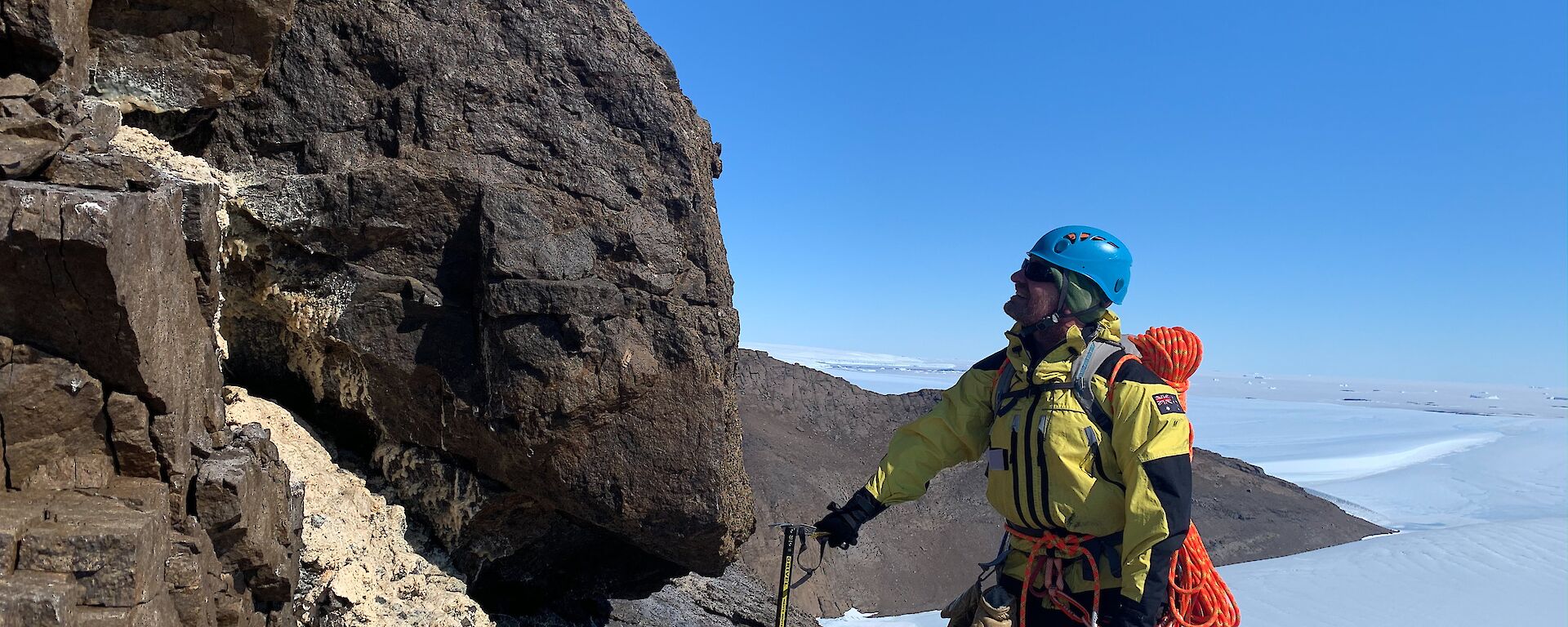 An climber with ropes and helmet stands atop a mountain looking at a mumiyo deposit.  Snow can be seen on the ground below.