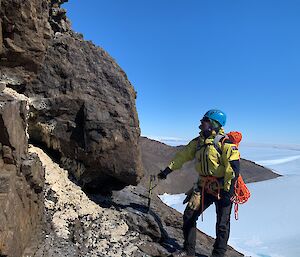 An climber with ropes and helmet stands atop a mountain looking at a mumiyo deposit.  Snow can be seen on the ground below.
