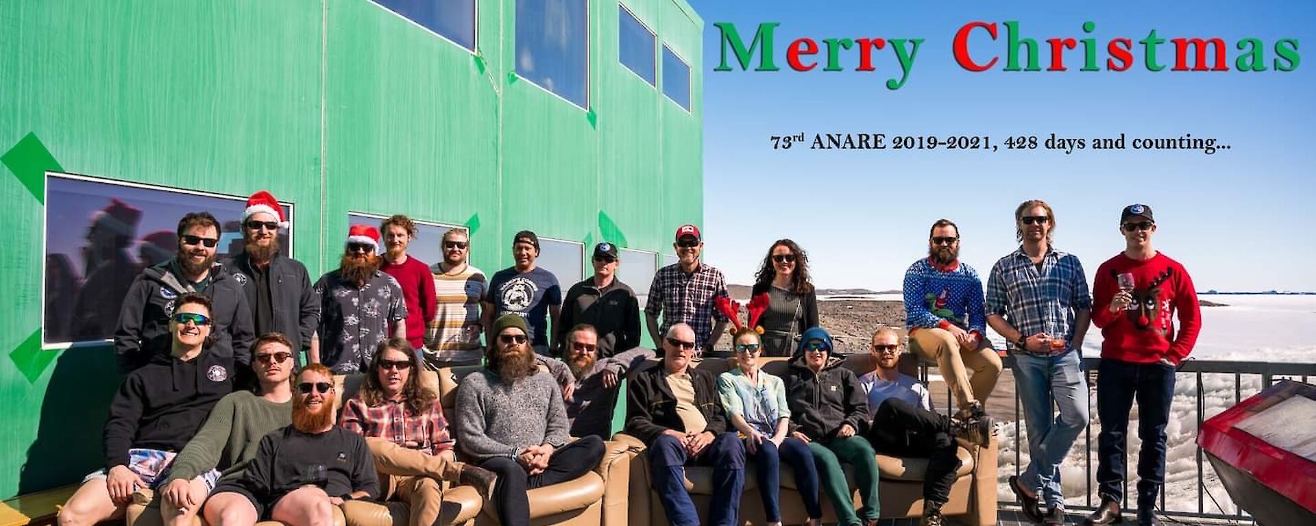 Expeditioners in a group photo on the outside deck, some with Santa hats on.  Text on photo reads 'From all of us here at Davis station, Merry Christmas.  73rd Anare 2019-20, 428 days and counting