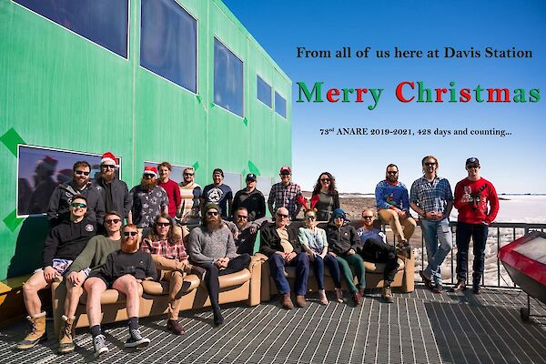 Expeditioners in a group photo on the outside deck, some with Santa hats on. Text on photo reads 'From all of us here at Davis station, Merry Christmas. 73rd Anare 2019-20, 428 days and counting