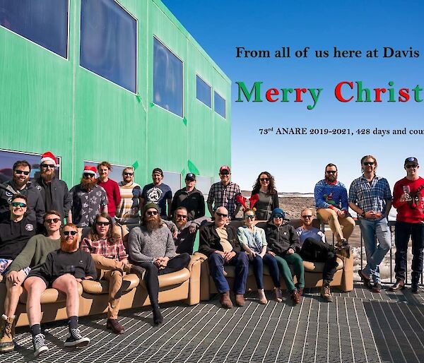 Expeditioners in a group photo on the outside deck, some with Santa hats on.  Text on photo reads 'From all of us here at Davis station, Merry Christmas.  73rd Anare 2019-20, 428 days and counting