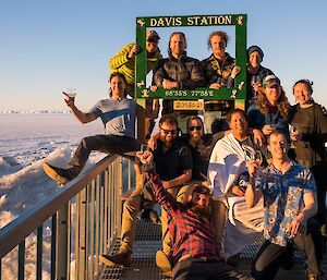 A group photo of expeditioners on the deck, holding drinks and smiling at midnight.  A large picture frame is being held up titled Davis Station with the co-ordinates, below which are the dates 2019-20-21.