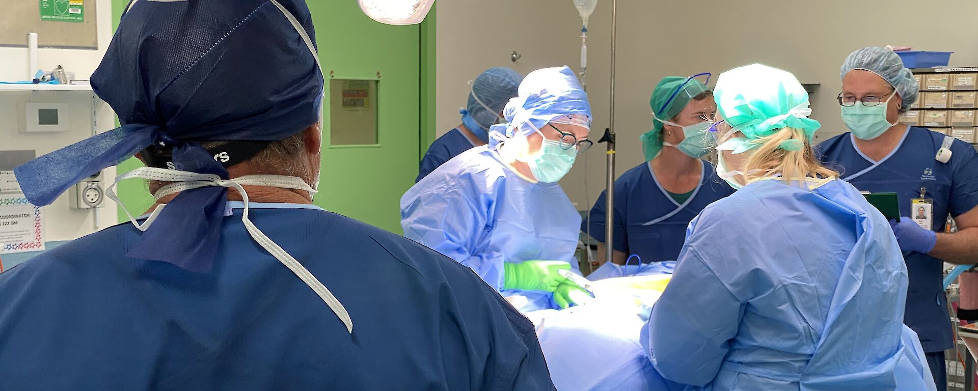 Back of man in surgical scrubs, watching several others in the operating theatre