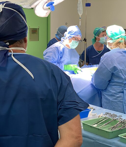 Back of man in surgical scrubs, watching several others in the operating theatre