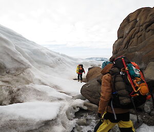 Two expeditioners with full packs walking between where the ice meets the rocks