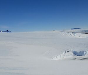 Icy landscape in foreground including ice cliffs with two mountain ranges on the horizon against a clear blue sky