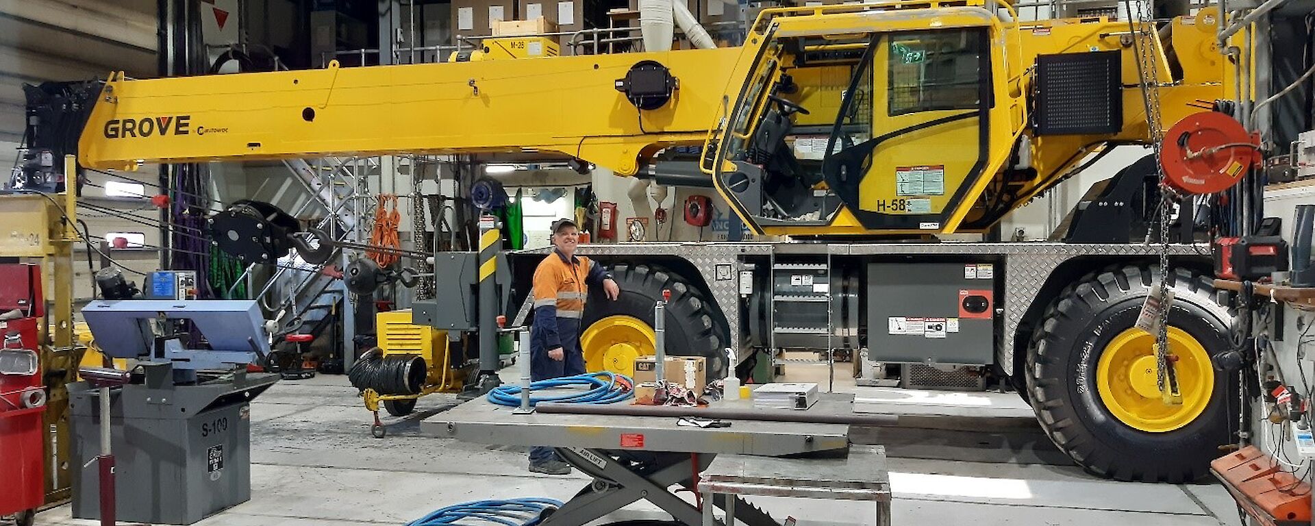 Large yellow crane inside a shed full of mechanical equipment.  Mechanic in hi-vis leans against the large wheel, smiling to camera.