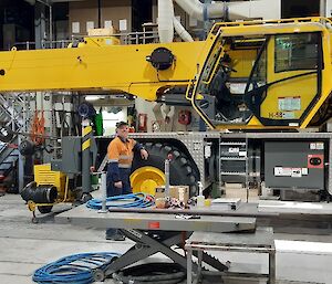 Large yellow crane inside a shed full of mechanical equipment.  Mechanic in hi-vis leans against the large wheel, smiling to camera.