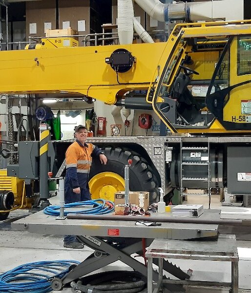Large yellow crane inside a shed full of mechanical equipment.  Mechanic in hi-vis leans against the large wheel, smiling to camera.