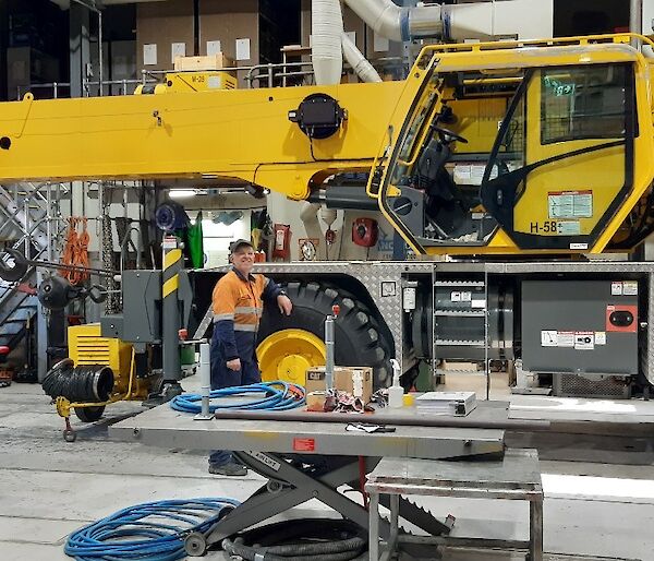 Large yellow crane inside a shed full of mechanical equipment. Mechanic in hi-vis leans against the large wheel, smiling to camera.