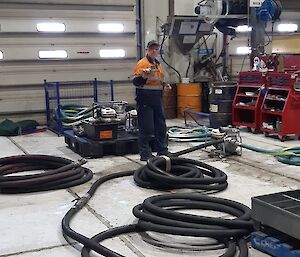 An expeditioner in hi-vis standing amongst coils of pipeline holding up a tool to camera