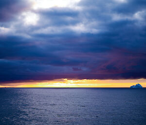 An orange sunset on the horizon with grey clouds above.  A floating iceberg can be seen in the distance.