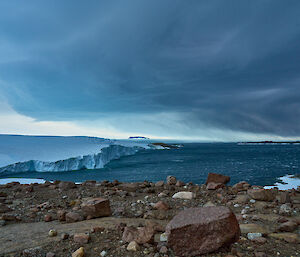 A rocky foreshore with an ice cliff falling to the sea.  Large grey storm clouds gather above.