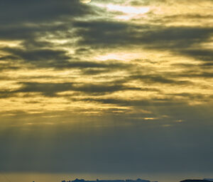 A yellow cloudy sky with sun rays shining down on to an icy landscape with a large iceberg in the distance