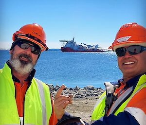 Two men in hi-vis and hard hats smiling to camera with the MPV Everest visible between them on the water