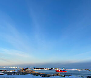 An orange supply vessel sits in the calm waters of the bay with a beautiful blue sky above and snow in the foreground