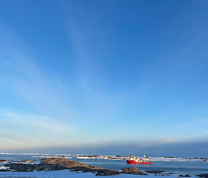 An orange supply vessel sits in the calm waters of the bay with a beautiful blue sky above and snow in the foreground