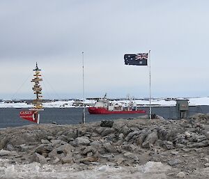 Australian flag on a flagpole and the Casey "directions" sign in foreground, ship in midground. Snow covered terrain in background.