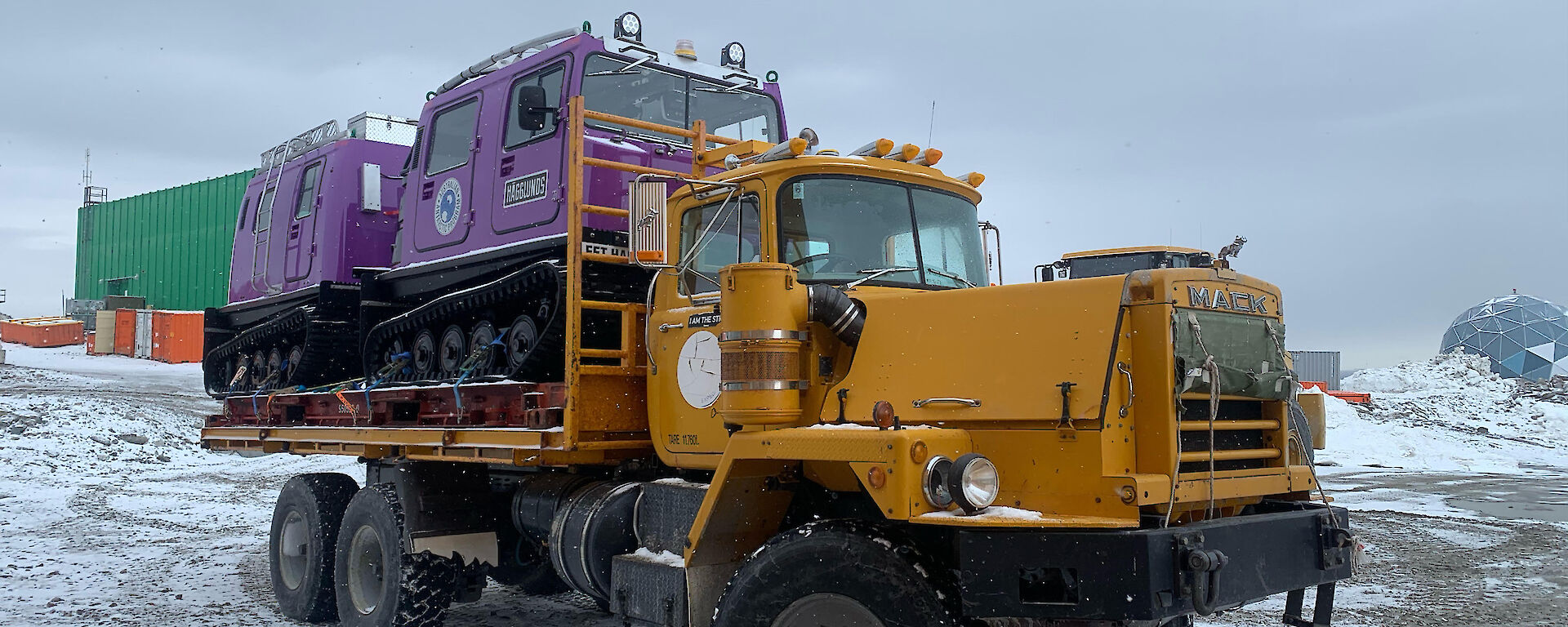 Purple Hägglunds vehicle on the back of a Mack truck.