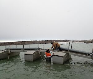 One expeditioner on a pontoon helping another who is waste deep in the water to clip the sections together