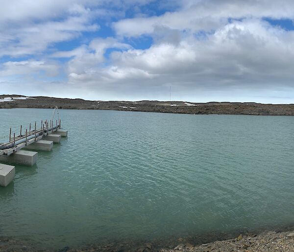 A panoramic of the melted water in the tarn with a short jetty and pipes leading in to it.