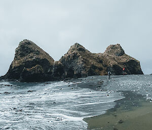 A large three peak rock in the background with the beach and waves on the shore in the foreground