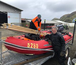 A man leaning against a dinghy on a trailer.  A man in hi-vis stands in the dingy strapping in some wood.