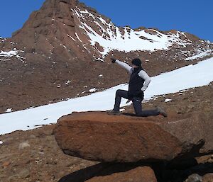 An expeditioner strikes a pose on a rock, kneeling and pointing to the distance
