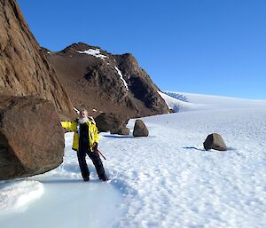 An expeditioner stands in the snow and ice with his hands on a huge rock at the side of him