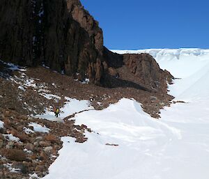 An expeditioner in the distance scrambling over some rocks with a moutain on one side and snow on the other