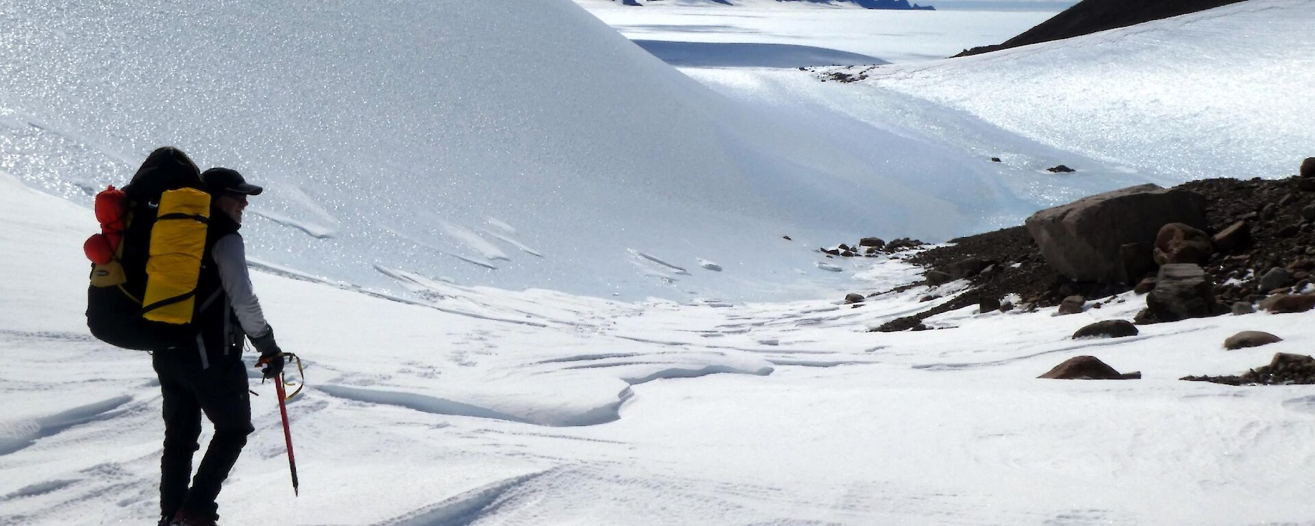 An expeditioner in backpack and hiking gear walking down a valley with mountain peaks visible on the horizon peeping out from the snowy ground