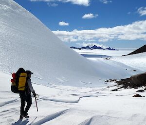 An expeditioner in backpack and hiking gear walking down a valley with mountain peaks visible on the horizon peeping out from the snowy ground