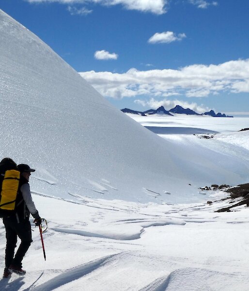 An expeditioner in backpack and hiking gear walking down a valley with mountain peaks visible on the horizon peeping out from the snowy ground