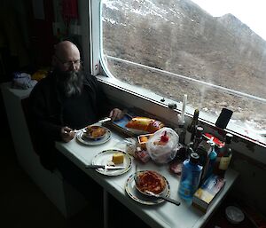 An expeditioner sits at a hut table with a meal in front of him and a window looking out to the rocky landscape