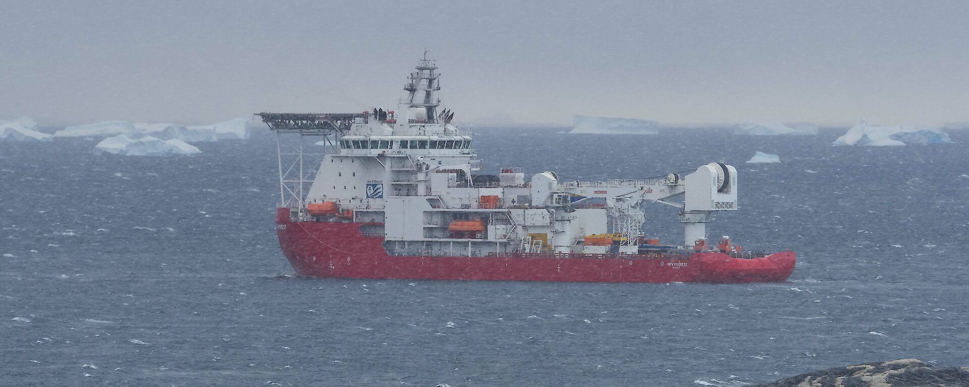 A red supply ship sailing away from the land with icebergs in the distance