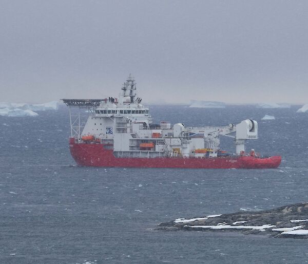 A red supply ship sailing away from the land with icebergs in the distance