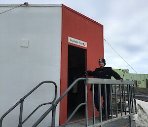 The postmaster standing at top of some stairs leading in to the red post office hut