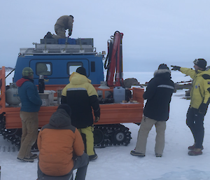 Four expeditioners standing round a Hagglund talking.  Another man kneels on the roof tying equipment and another rests on an Eski