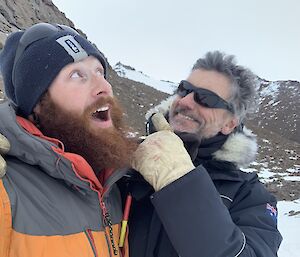 Close up of two expeditioners, one has his arm around the other and is pointing behind him, the other looks upwards