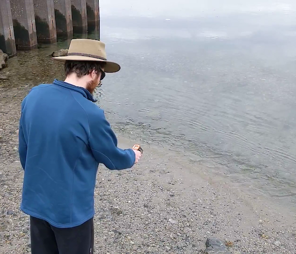 An expeditioner stands at the shoreline and looks at a thermometer