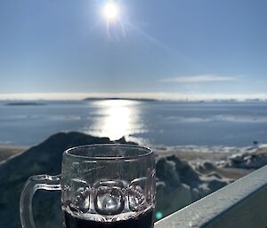 A pint glass sitting on a balcony railing with the sun reflected of the sea in the distance