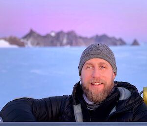 A portrait of an expeditioner smiling to camera with a woollen beanie on with a pink sunset, sea ice and mountians behind.